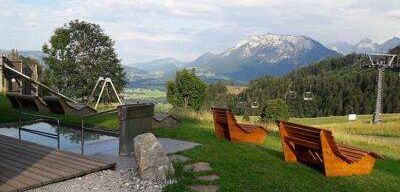 Eine ruhige Außenfläche mit Holzbänken und einem Wasserelement. Berge und Bäume in der Ferne. Ein Skilift ist auf der rechten Seite sichtbar.