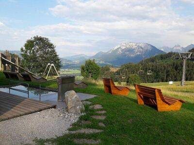 Eine ruhige Außenfläche mit Holzbänken und einem Wasserelement. Berge und Bäume in der Ferne. Ein Skilift ist auf der rechten Seite sichtbar.