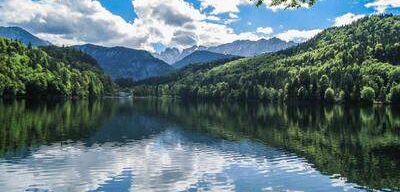 Ein ruhiger See mit klarem blauem Wasser spiegelt den blauen Himmel und flauschige weiße Wolken wider. Dichte grüne Berge umgeben den See und schaffen eine malerische Naturszene.