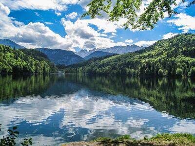 Ein ruhiger See mit klarem blauem Wasser spiegelt den blauen Himmel und flauschige weiße Wolken wider. Dichte grüne Berge umgeben den See und schaffen eine malerische Naturszene.