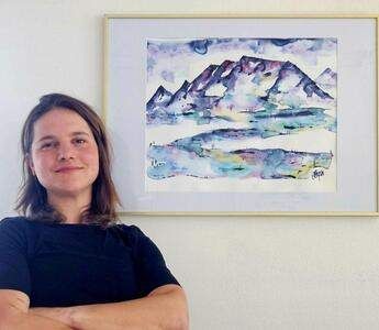 Eine Frau steht mit verschränkten Armen vor einem Aquarellbild von Bergen. Das Gemälde ist in einem goldenen Rahmen gefasst.