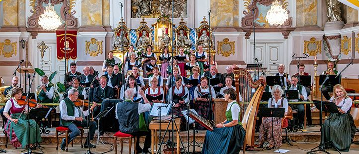 Ein Chor und ein Orchester treten in einem großen, reich verzierten Saal auf. Musiker spielen verschiedene Instrumente, während sie auf der Bühne sitzen.