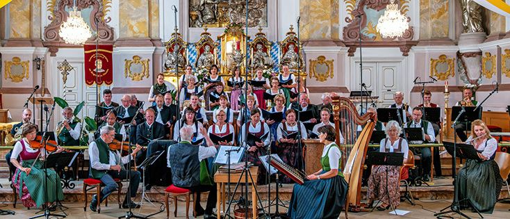 Ein Chor und ein Orchester spielen in einer barocken Kirche, geschmückt mit goldenen Dekorationen und hängenden Bannern. Die Musiker spielen eine Vielzahl von Instrumenten, darunter Celli, Harfen und Holzblasinstrumente.