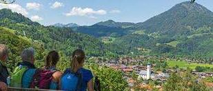 Vier Wanderer, mit Rucksäcken, sitzen auf einer Bank mit Blick auf ein Tal mit einem Dorf und Bergen im Hintergrund.
