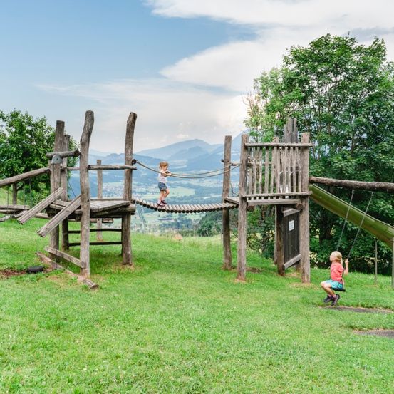 Zwei Kinder spielen an einem sonnigen Tag auf einem Holzspielplatz mit einer Rutsche und einer Seilbrücke.
