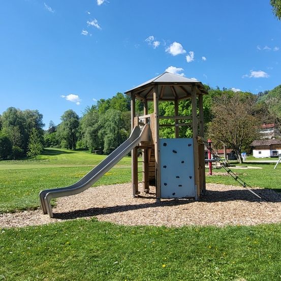 Ein Spielbereich mit Rutsche und Kletterwand in einem Park, umgeben von Gras und Bäumen, unter einem blauen Himmel mit Wolken.