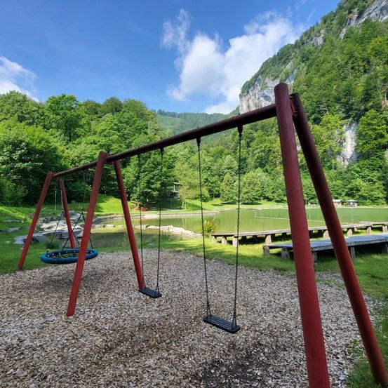 Ein Outdoor-Spielplatz mit Schaukeln und einem Reifenschaukel in einer grünen Umgebung mit einem See und einem Felsen im Hintergrund.