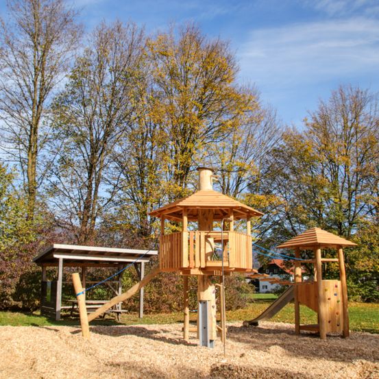 Ein holzspielhaus mit rutsche und kletterwand befindet sich in einem park mit herbstbäumen.