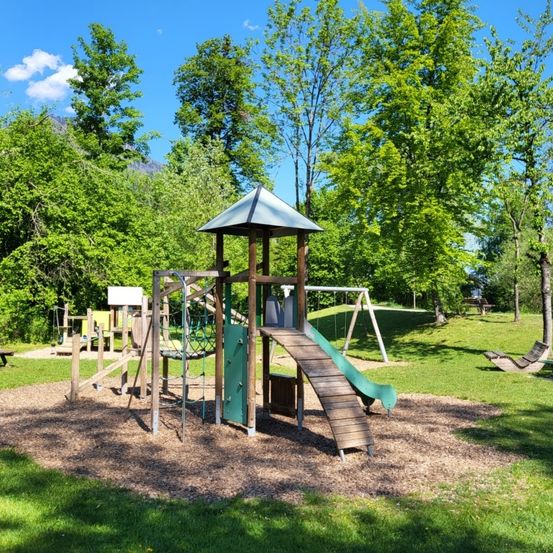 Ein Spielplatz mit Rutsche, Kletterwand und Schaukeln. Grüner Rasen und Bäume umgeben das Gelände.