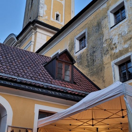 Ein gelbes Gebäude mit Turm, Fenstern und rotem Dach steht vor einem weißen Zelt. Das Zelt hat Metallstangen und zwei leuchtende Laternen.