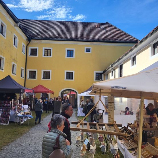 Ein Outdoor-Markt mit mehreren Ständen findet vor einem gelben Gebäude mit vielen Fenstern an einem sonnigen Tag statt. Menschen schauen sich die Artikel an den Ständen an.