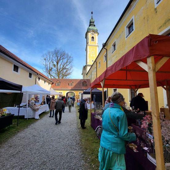 Ein Freiluftmarkt mit mehreren Ständen unter Zelten, umgeben von gelben Gebäuden. Menschen gehen vorbei und schauen sich die ausgestellten Artikel an. Ein Turm ist im Hintergrund sichtbar.