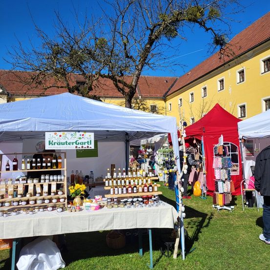 Ein Freiluftmarkt mit Ständen unter Zelten vor einem gelben Gebäude. Ein Zelt zeigt Gläser und Flaschen mit Produkten. Eine Person steht im Hintergrund.