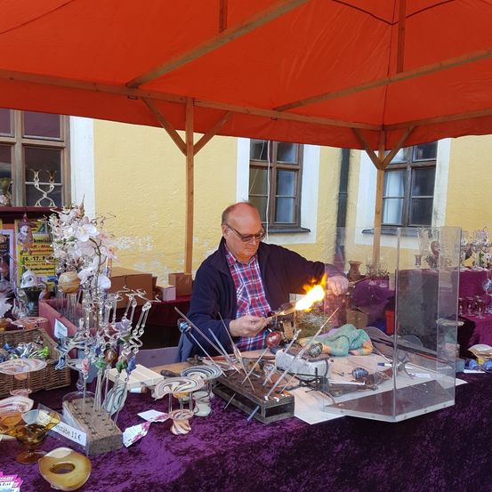 Ein älterer Mann mit Brille arbeitet an einem Glasblasprojekt auf einem Outdoor-Markt. Gläserne Gegenstände sind auf dem Tisch ausgestellt.