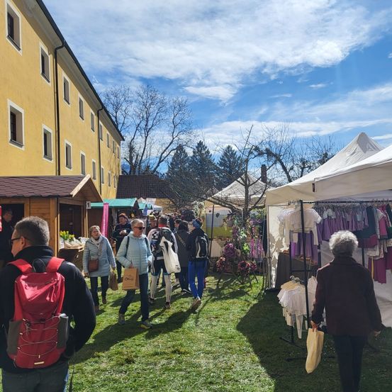 Menschen spazieren auf einem Markt mit Ständen unter weißen Zelten, in der Nähe eines gelben Gebäudes mit vielen Fenstern, an einem sonnigen Tag.