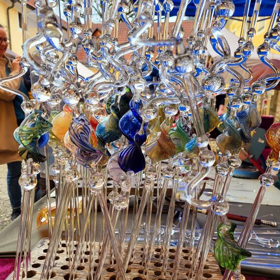 Eine Ausstellung von farbenfrohen Glaskunstwerken, darunter Fische, Blumen und Blätter, auf einem Holzständer angeordnet. Sie befinden sich unter einem blauen Zelt.