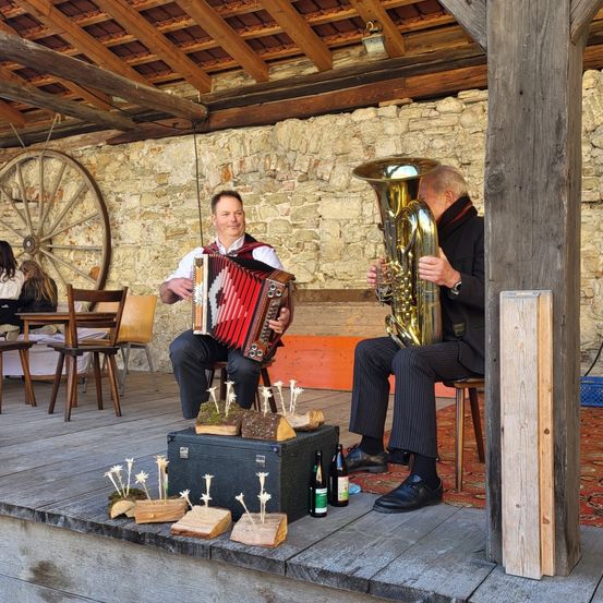 Zwei Männer spielen Musik auf einer Bühne mit einem Hintergrund aus Steinmauern. Einer spielt Tuba, der andere Akkordeon. Sie sitzen auf Stühlen, Flaschen und Pflanzen sind in der Nähe.