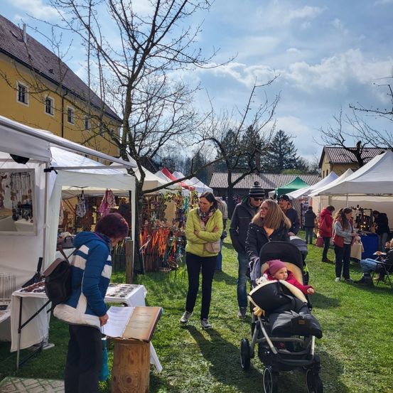 Mehrere Menschen gehen auf einem Markt mit Ständen umher. Eine Frau liest einen Prospekt. Eine Frau mit einem Kinderwagen schaut auf ein Kind. Andere Menschen gehen herum und schauen auf die Stände.
