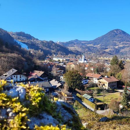 Luftaufnahme eines Dorfes in einer Bergregion mit Häusern, Bäumen und einer Kirche. Berge im Hintergrund unter einem klaren blauen Himmel.