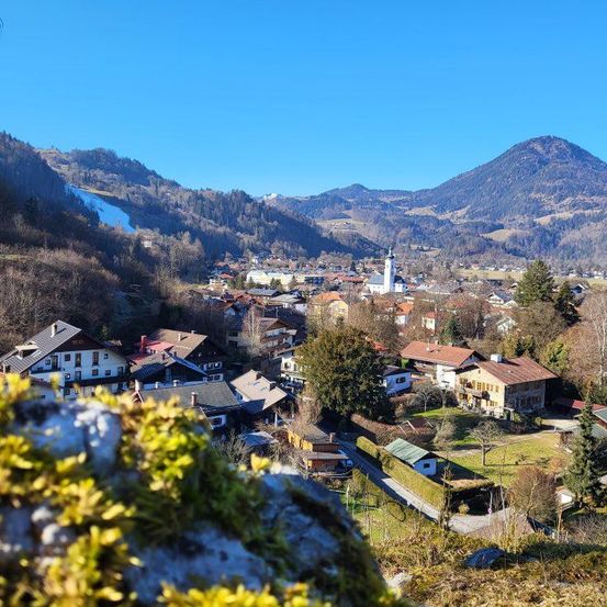 Luftaufnahme eines Dorfes in einer Bergregion mit Häusern, Bäumen und einer Kirche. Berge im Hintergrund unter einem klaren blauen Himmel.