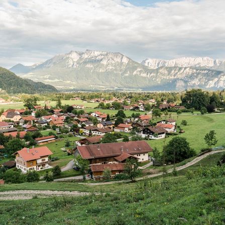 Ein Dorf in einem Tal, umgeben von Bergen, mit Häusern und Bäumen unter einem bewölkten Himmel.