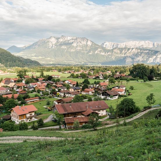 Ein Dorf in einem Tal, umgeben von Bergen, mit Häusern und Bäumen unter einem bewölkten Himmel.