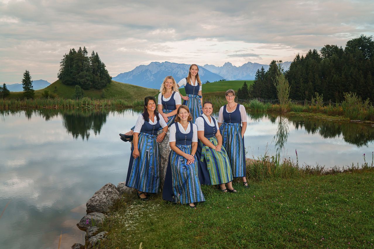 Sechs Frauen in traditioneller Kleidung posieren für ein Foto am See mit Bergen im Hintergrund.
