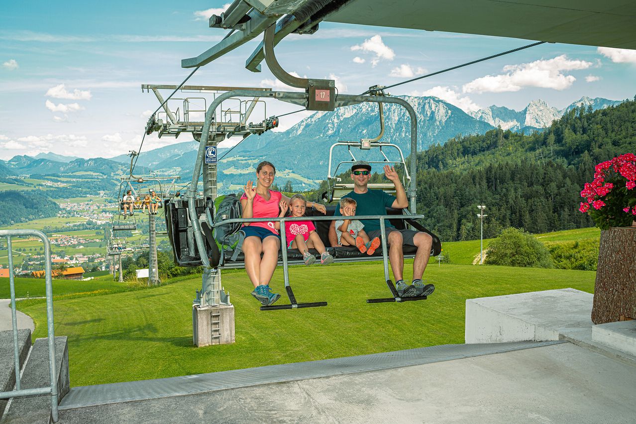 Eine vierköpfige Familie fährt mit einer Skilift in den Bergen mit einer malerischen Aussicht auf grüne Berge und Bäume.