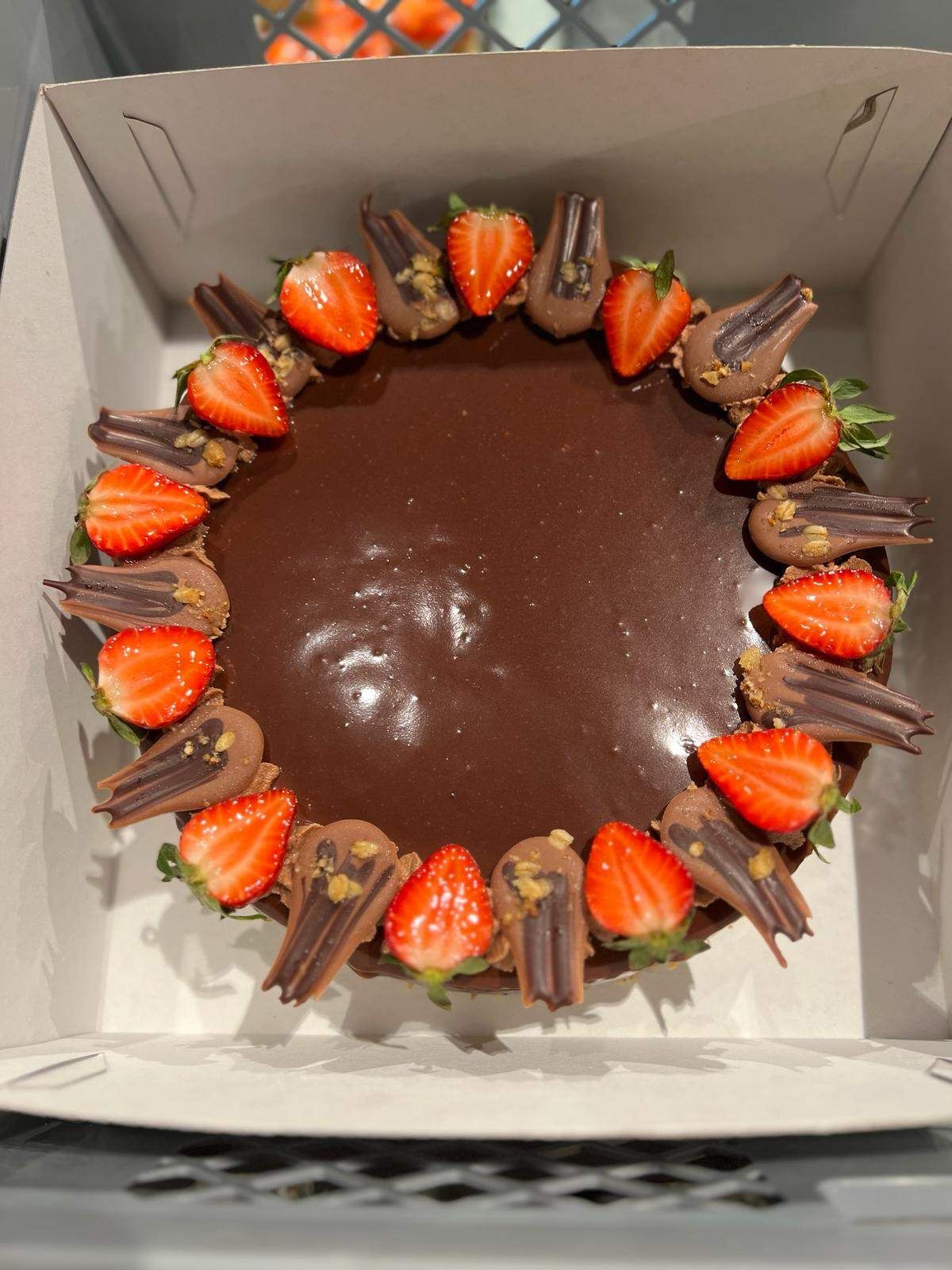 Ein runder Schokoladenkuchen mit geschnittenen Erdbeeren und Schokoladenraspeln ist in einer Box platziert. Der Kuchen hat eine glatte, glänzende Schokoladenschicht.