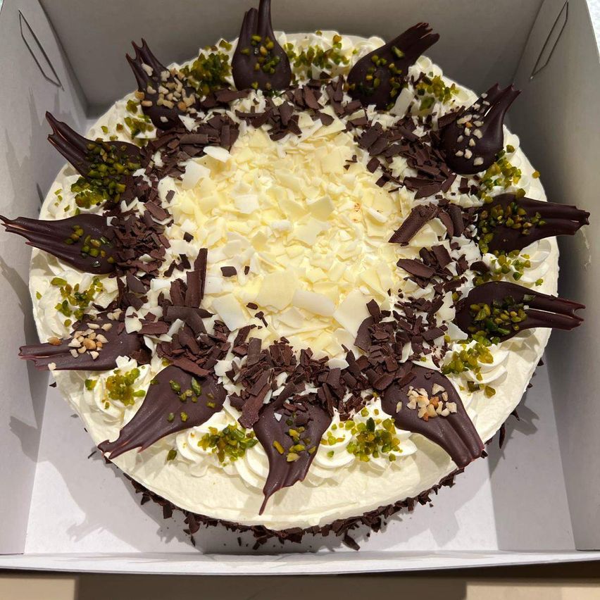 Ein runder Kuchen mit weißer Cremebasis, belegt mit Schokoladenlocken, Pistazien und Kokosraspeln, in einer weißen Schachtel.