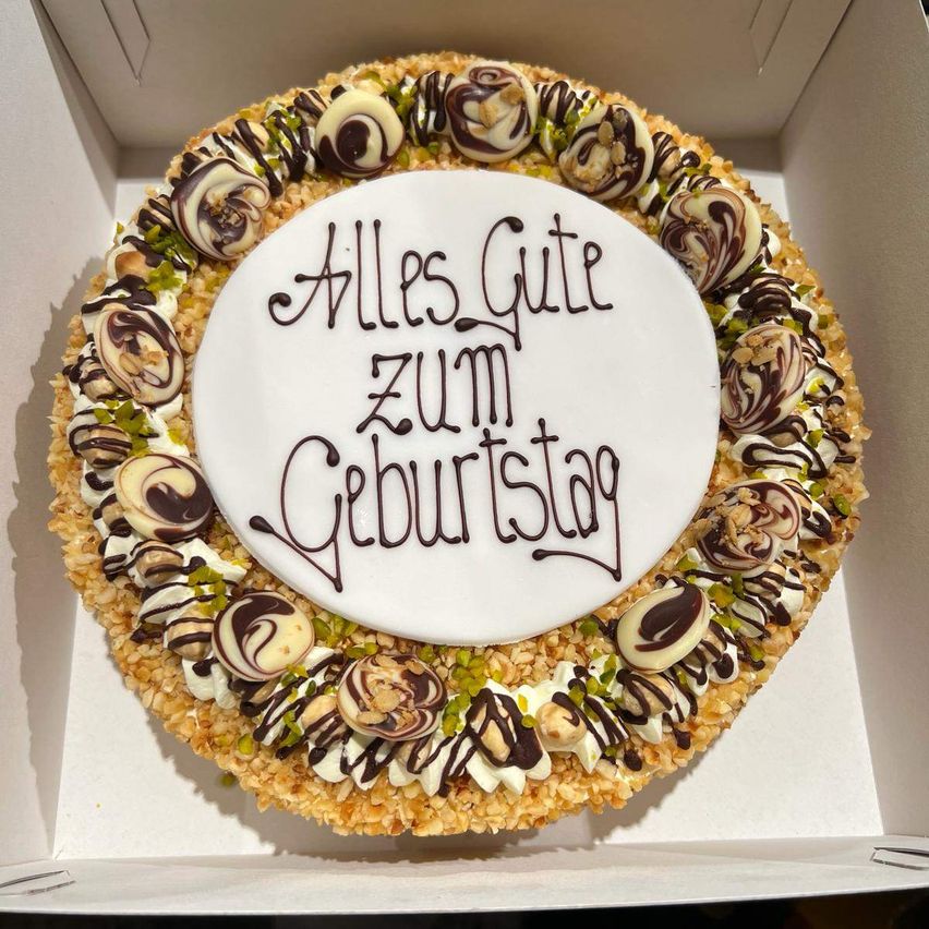 Ein Kuchen in einer Box mit einer weißen runden Tafel, dekoriert mit Schokoladenwirbeln und Nüssen, mit den Worten 'Alles Gute zum Geburtstag' in schwarzer Schrift geschrieben.
