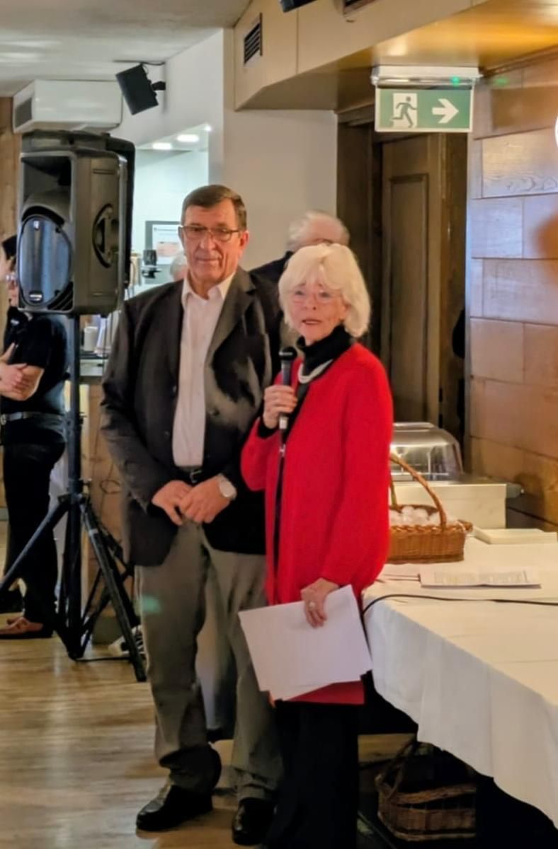 Ein älterer Mann und eine ältere Frau stehen neben einem Tisch mit einem Mikrofon. Sie halten Papiere und lächeln. Ein Lautsprecher und ein Korb stehen hinter ihnen.