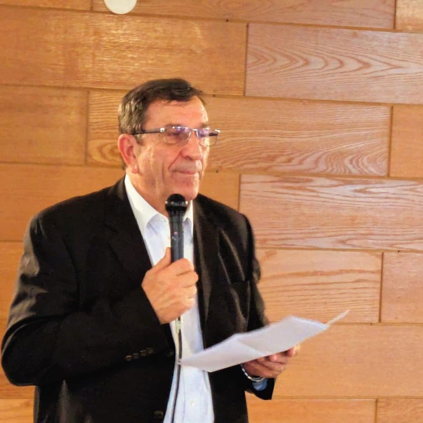 Ein älterer Mann mit Brille und schwarzem Anzug spricht in ein Mikrofon und hält ein Blatt Papier in der Hand. Er scheint eine Rede zu halten. Der Hintergrund ist eine Holzwand.