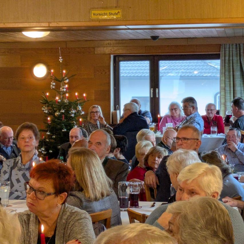 Eine Gruppe älterer Menschen sitzt um Tische in einem Esszimmer herum. Ein mit Kerzen bestückter Weihnachtsbaum steht in der Mitte. Der Raum hat Holzwände und ein großes Fenster mit Vorhängen.