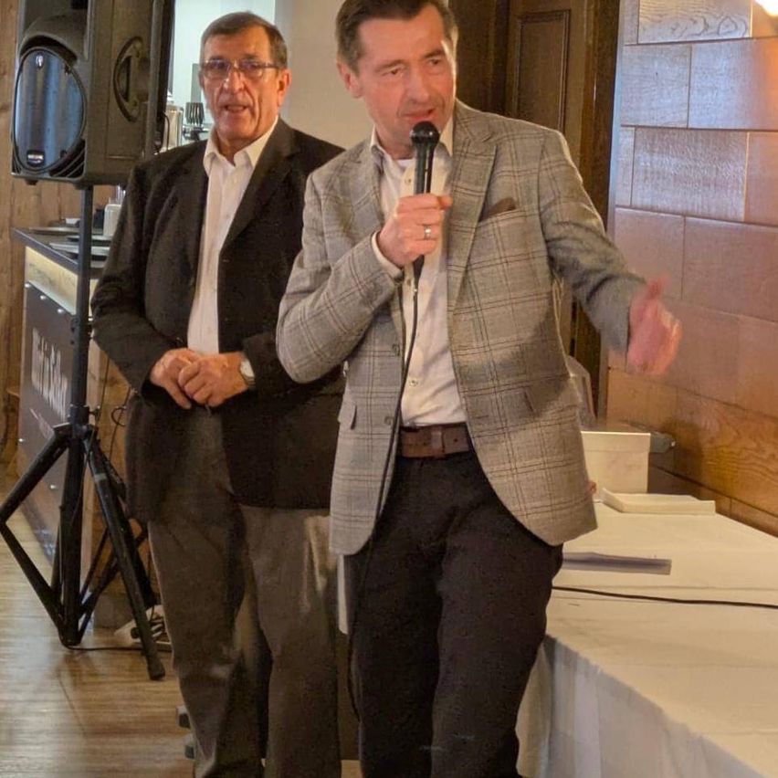 Ein Mann spricht in ein Mikrofon, während ein anderer Mann hinter ihm steht und zuhört. Die Umgebung scheint ein Konferenz- oder Besprechungsraum zu sein.