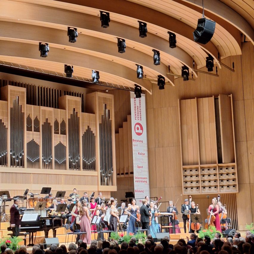 Ein Konzert findet in einem großen Konzertsaal statt. Ein Orchester spielt auf der Bühne mit einem Flügel, Violinen, Celli und einer großen Orgel im Hintergrund. Das Publikum sitzt in den vorderen Reihen.