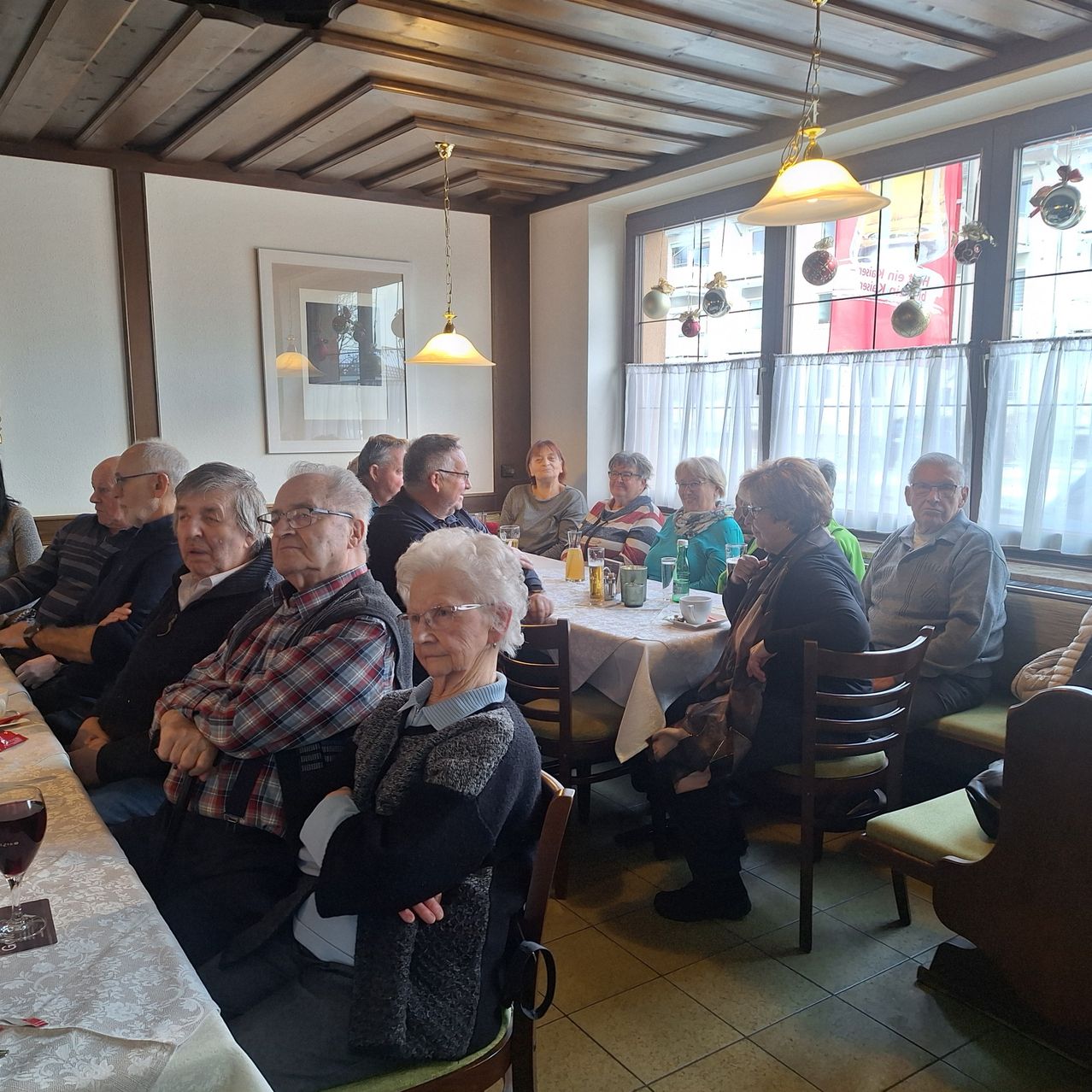 Several elderly people sit at tables in a restaurant, with drinks and food on the tables. The room has white walls, wooden ceiling, and windows with white curtains.