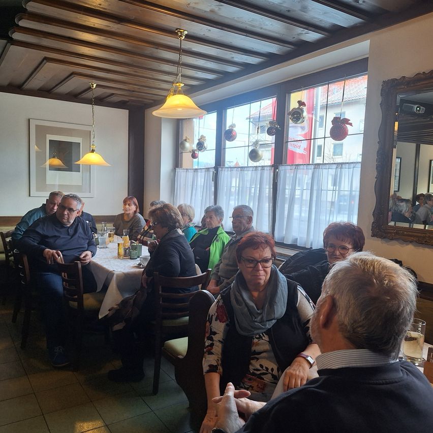 A group of people are sitting around a table in a restaurant, with a woman wearing glasses in the foreground. Behind her, others are seated, some wearing scarves. A window with curtains is visible in the background.