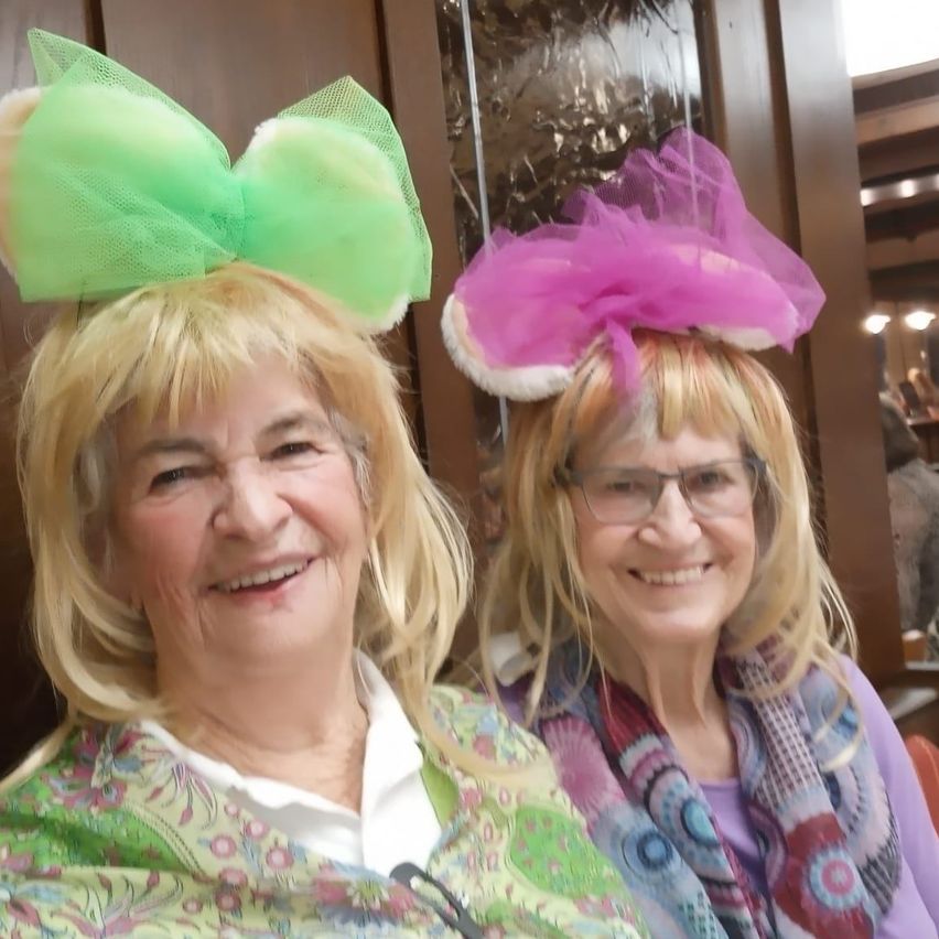 Zwei Frauen mit blonden Haaren, die bunte Kleidung tragen und lächeln. Eine hat eine grüne Haarschleife, die andere eine rosa. Sie befinden sich in einer Innenumgebung.