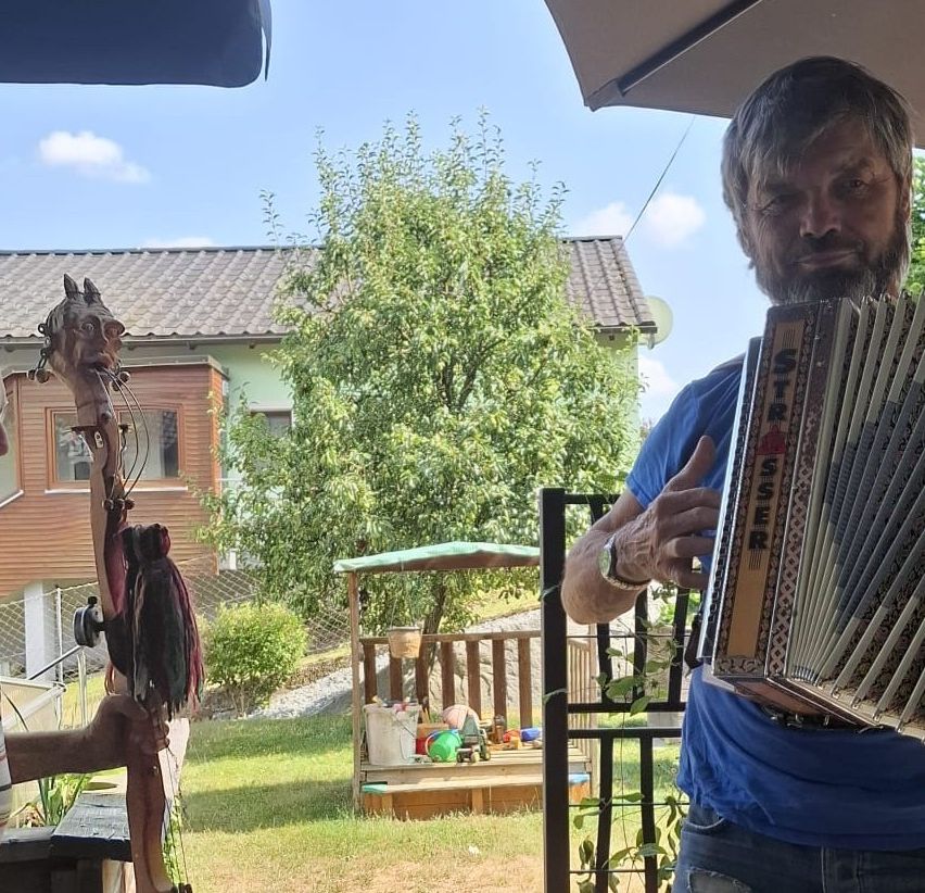 Ein Mann spielt Akkordeon unter einem Regenschirm im Garten mit einem Holzhaus und einem Baum im Hintergrund.