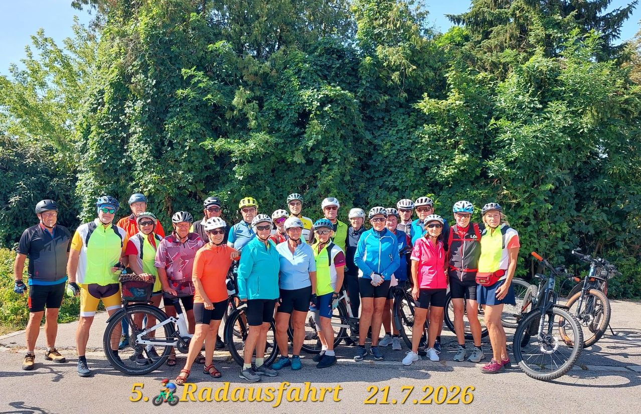 Eine Gruppe von Radfahrern in verschiedenfarbigen Trikots und Helmen posiert für ein Foto an einem sonnigen Tag vor einem grünen Hintergrund. Das Bild enthält einen Datumsstempel vom 21.7.2026.