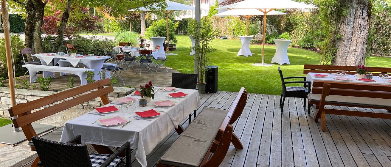 Ein Gartenrestaurant mit Holztischen und -bänken, weißen Tischdecken und roten Servietten. Der Bereich ist von weißen Sonnenschirmen beschattet und von üppigem Grün umgeben.