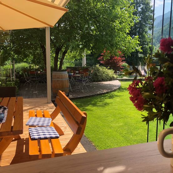 Eine Holzbank mit Kissen steht unter einem Terrassenschirm. Ein Tisch mit einer Tasse ist in der Nähe. Ein großer Baum und ein Fass sind im Hintergrund.