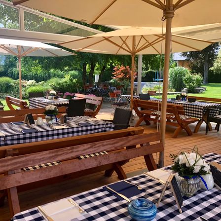 Ein Außenrestaurant mit Holztischen und -bänken unter großen Sonnenschirmen. Jeder Tisch hat eine karierte Tischdecke, eine Blumenvase und Besteck. Die Umgebung ist üppig mit Grün und Bäumen.