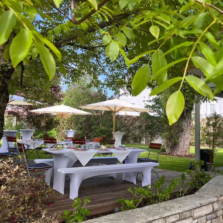 Eine Gartenparty mit weißen Tischdecken, Stühlen und Sonnenschirmen unter einem großen Baum. Der Bereich ist von viel Grün umgeben, mit Blick auf ein Haus in der Ferne.