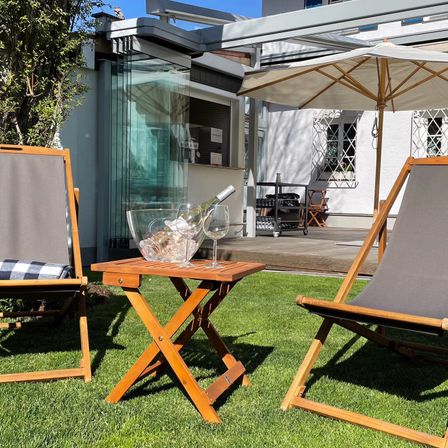 Zwei Holzstühle stehen auf einem Rasen, ein Tisch mit Wein und Weingläsern dazwischen. Im Hintergrund befinden sich ein Schirm und ein Haus.