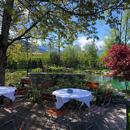 Ein Gartenrestaurant mit Tischen und Stühlen unter Bäumen, einem Teich und Bergen in der Ferne.