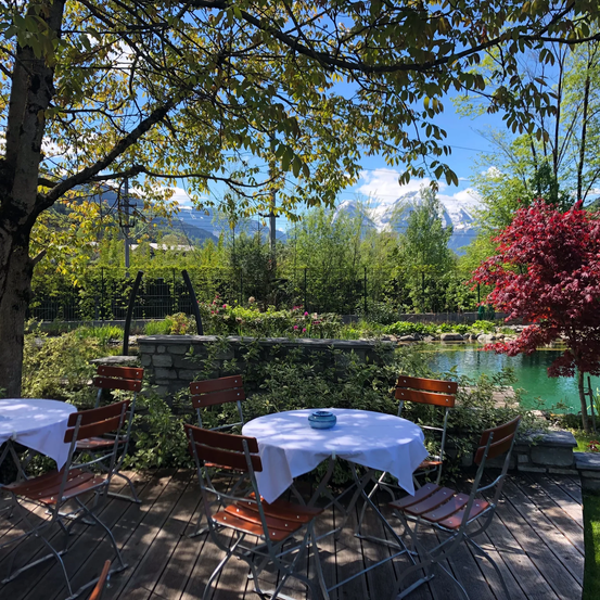 Ein Gartenrestaurant mit Tischen und Stühlen unter Bäumen, einem Teich und Bergen in der Ferne.