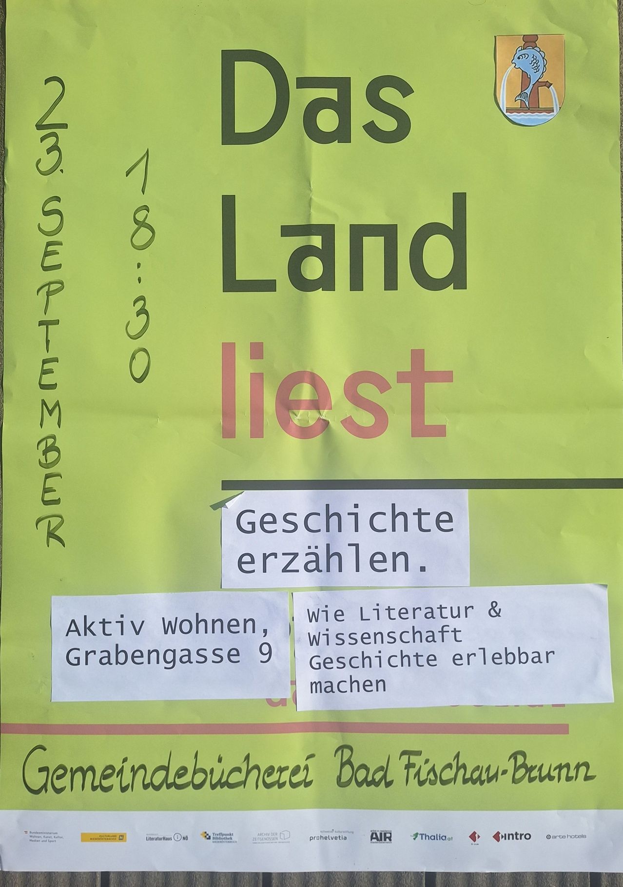 Ein grünes Plakat hat fette schwarzen und roten Text, der 'Das Land liest' lautet. Darunter steht 'Geschichte erzählen'. Ein weißer Karton sagt 'Aktiv wohnen, Grabengasse 9'. Ein weiterer weißer Karton sagt 'wie Literatur & Wissenschaft Geschichte erlebbar machen'.