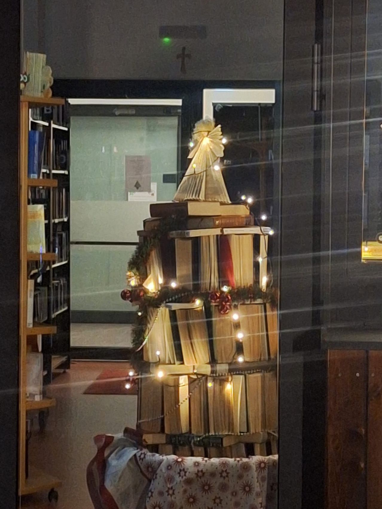 Ein dekorativer Weihnachtsbaum aus Büchern ist in einer Bibliothek ausgestellt. Er ist mit Lichtern und Ornamenten geschmückt.